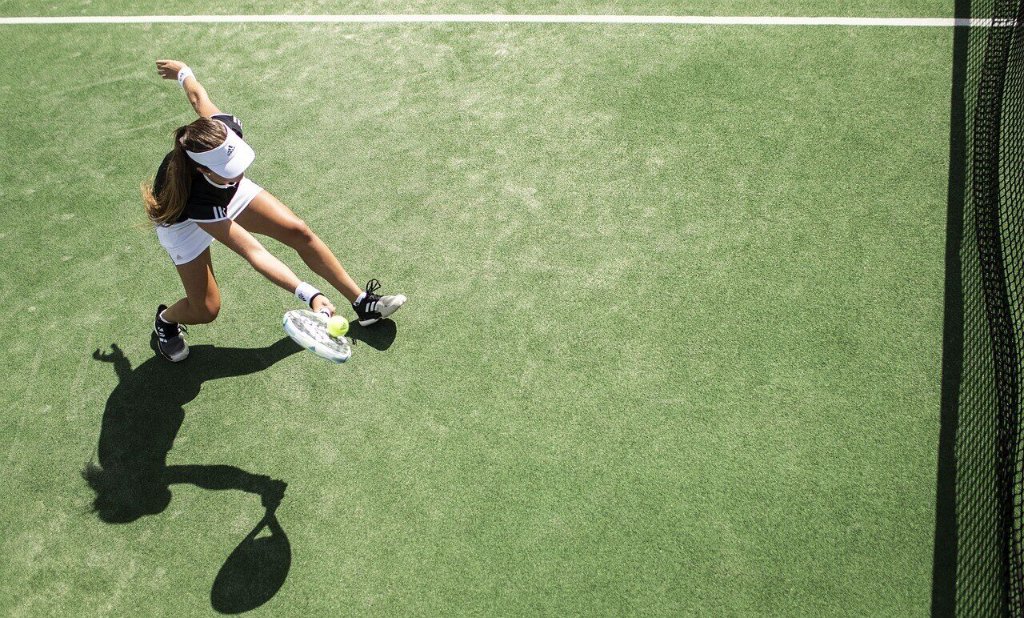 The Rise of Emma Raducanu: image of Overhead view of a tennis player on a green court, wearing a visor and athletic outfit, lunging to hit the ball near the net with a shadow cast prominently on the ground.