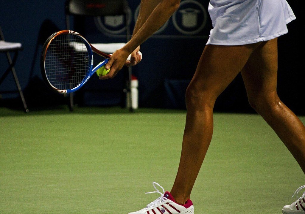 Close-up of a tennis player's legs and hands preparing to serve, holding a tennis ball and racket on an indoor court with blurred chairs and equipment in the background.